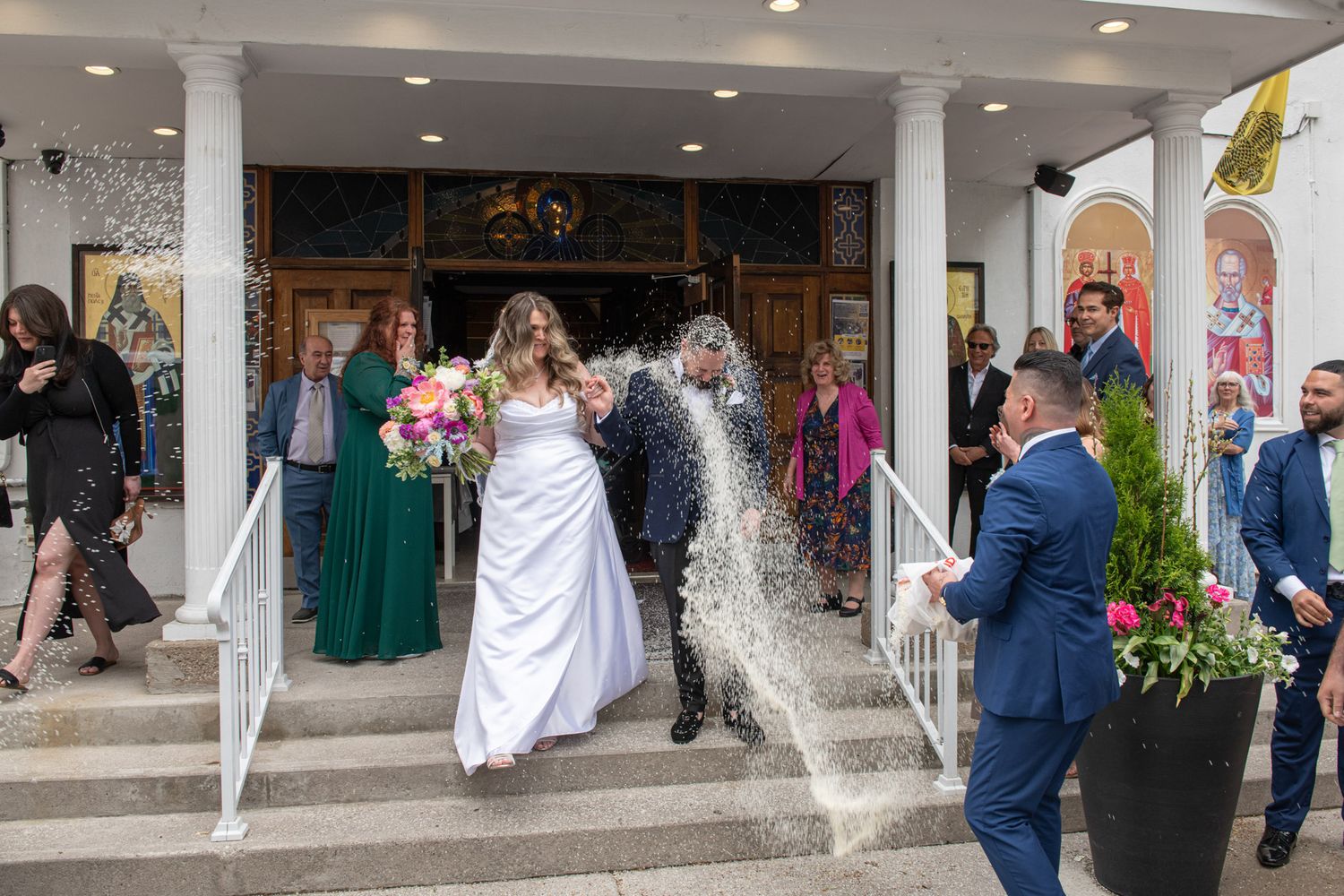 Bride and groom exit church greeted by rice toss from wedding guests on steps.