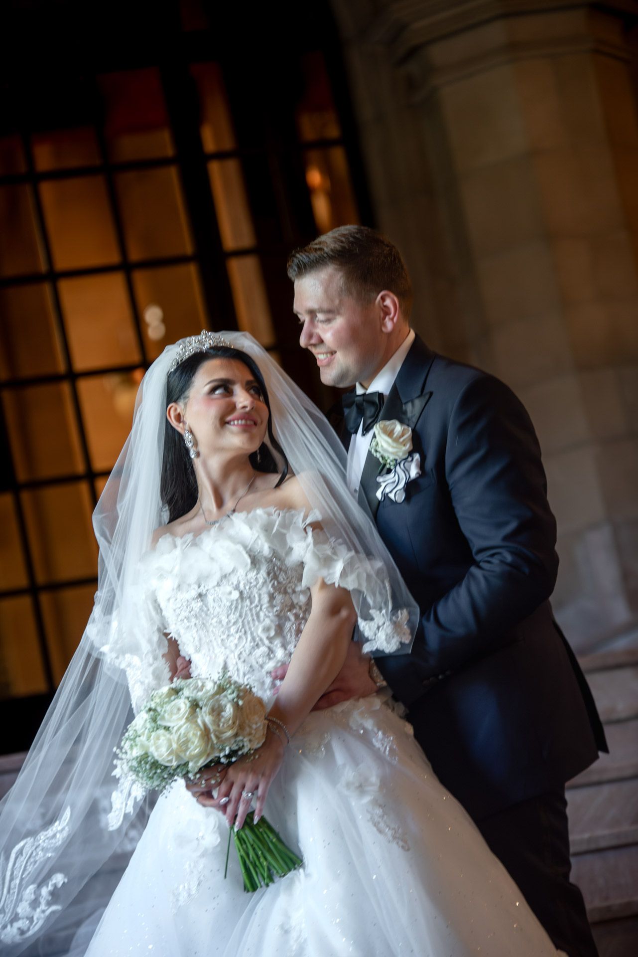 Elegant bride in lace gown and groom in tuxedo share tender moment at night.