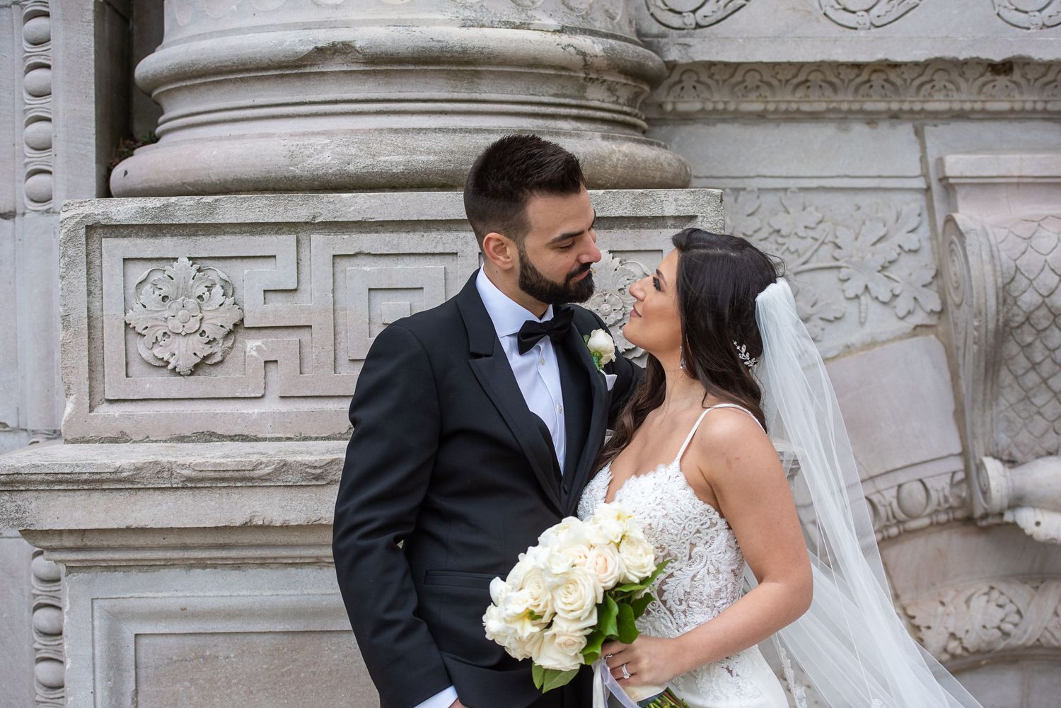 Bride holding white bouquet and groom in tuxedo posing by ornate stone architecture at Guildwood Park, Toronto.