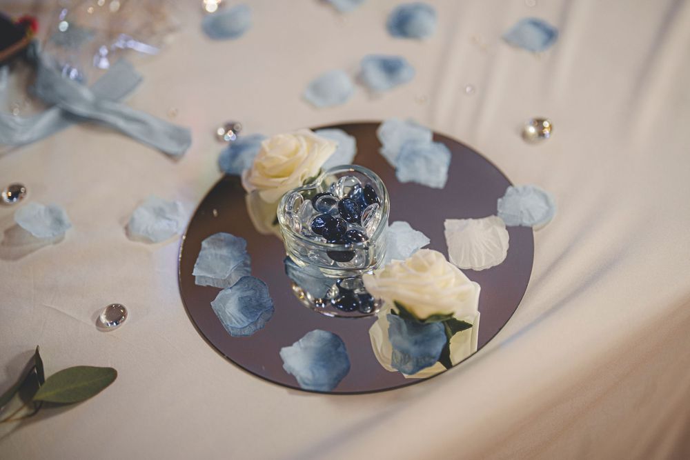 Wedding table decoration with white roses and blue petals on mirrored centerpiece.