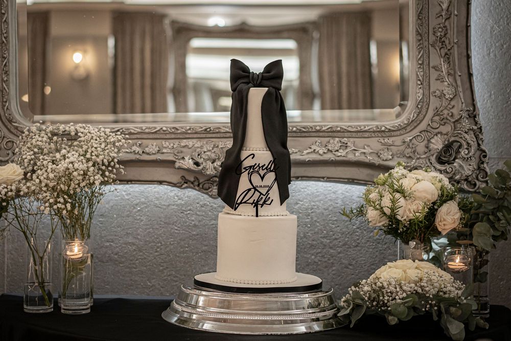 Elegant white wedding cake with bow tie topper displayed in front of ornate mirror.