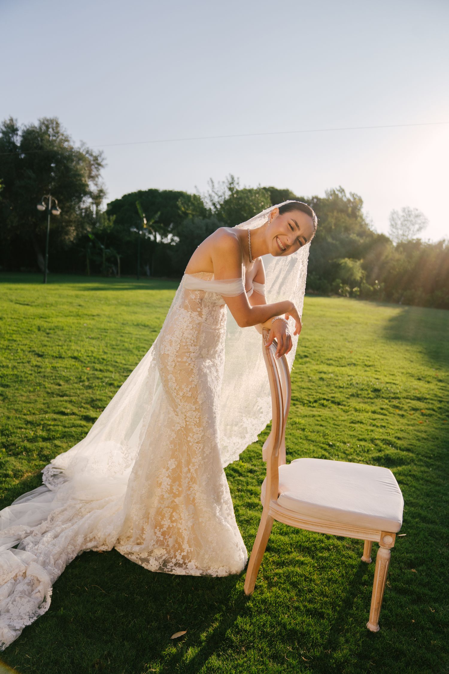 A bride in a lace wedding dress poses gracefully with a wooden chair on a sunlit grassy hill at golden hour.