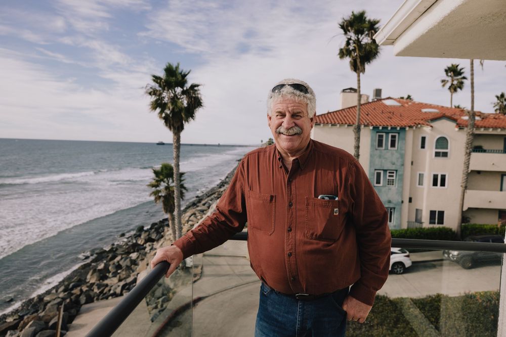 Older man smiling on oceanfront balcony with palm trees and luxury home in background.