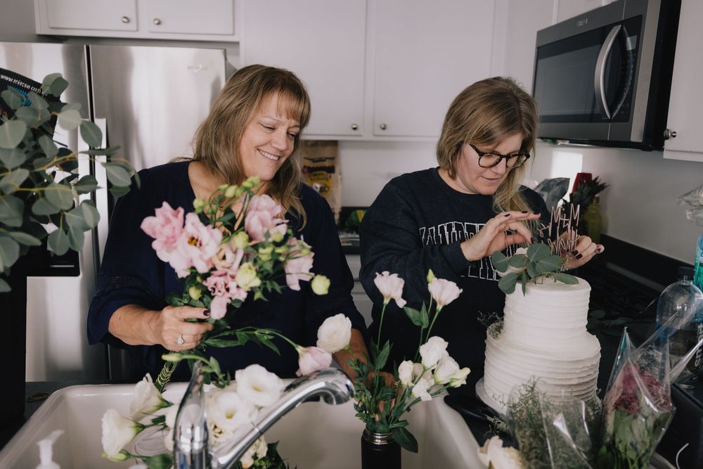 Two women arranging floral centerpieces and decorating a wedding cake in a kitchen.