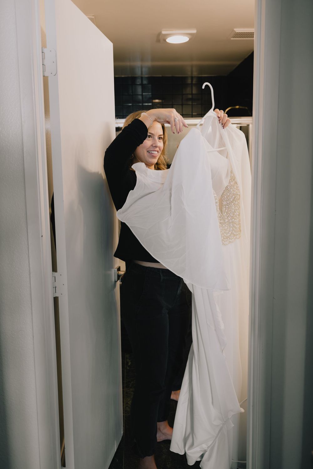 Blonde bride smiling while holding her beaded wedding gown in a dressing room.