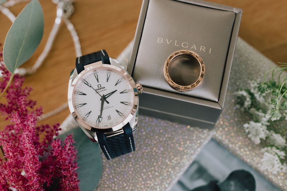 Omega watch with black leather strap beside a Bvlgari ring in box, surrounded by pink and white flowers.