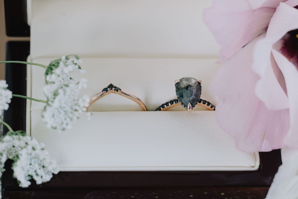 Rose gold engagement ring with teal pear-shaped gemstone and black diamond accents displayed in a white ring box.