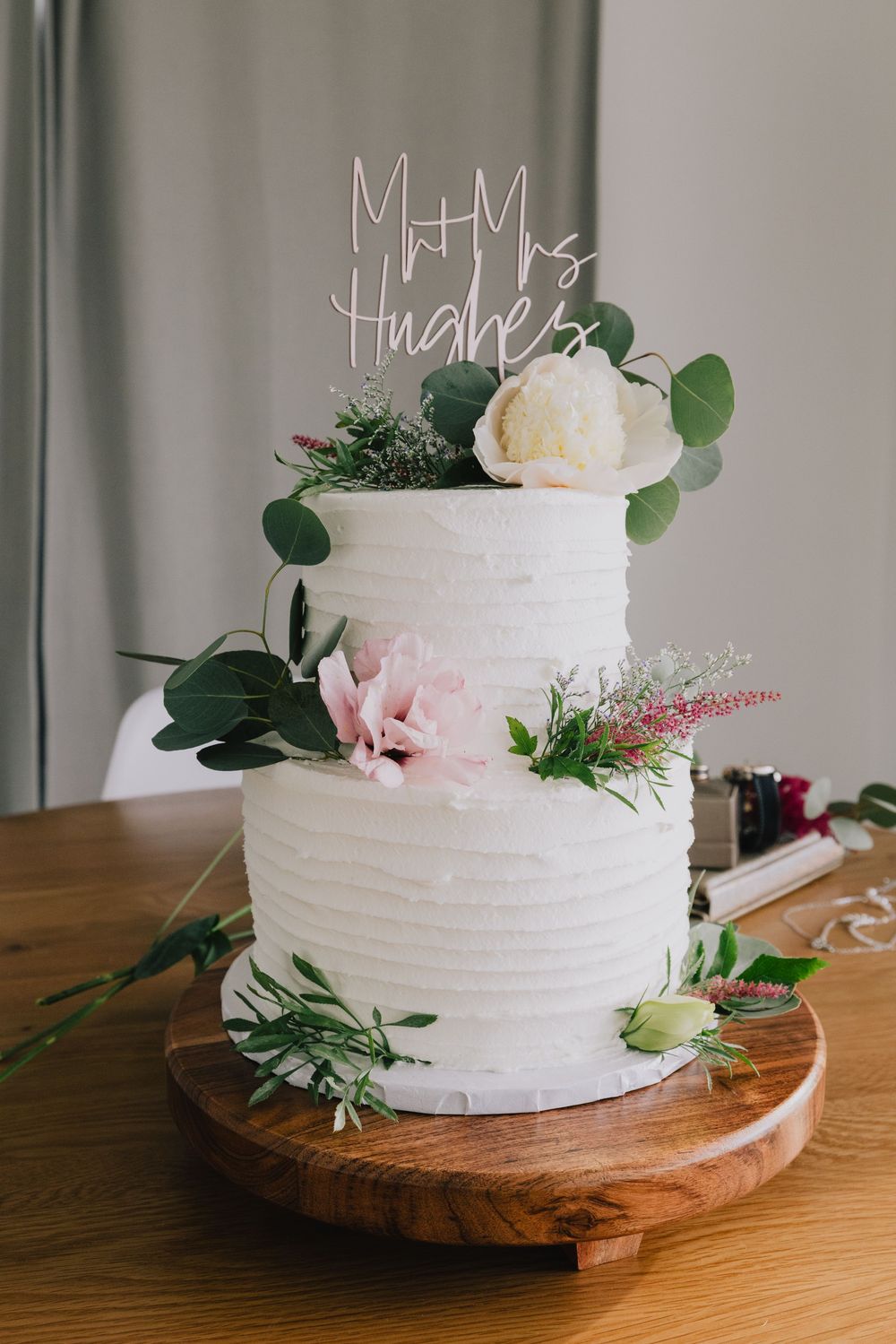 Two-tier white wedding cake with floral decorations and Mr & Mrs Hughes cake topper on wooden stand.