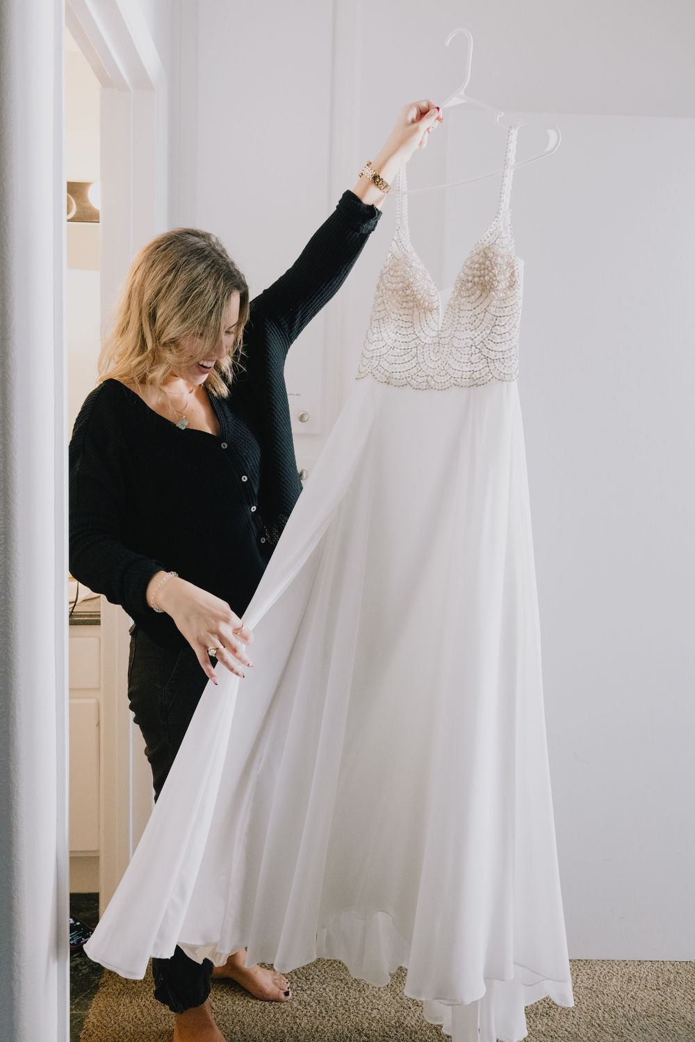 Blonde woman in black outfit holding up elegant beaded white wedding dress on hanger.