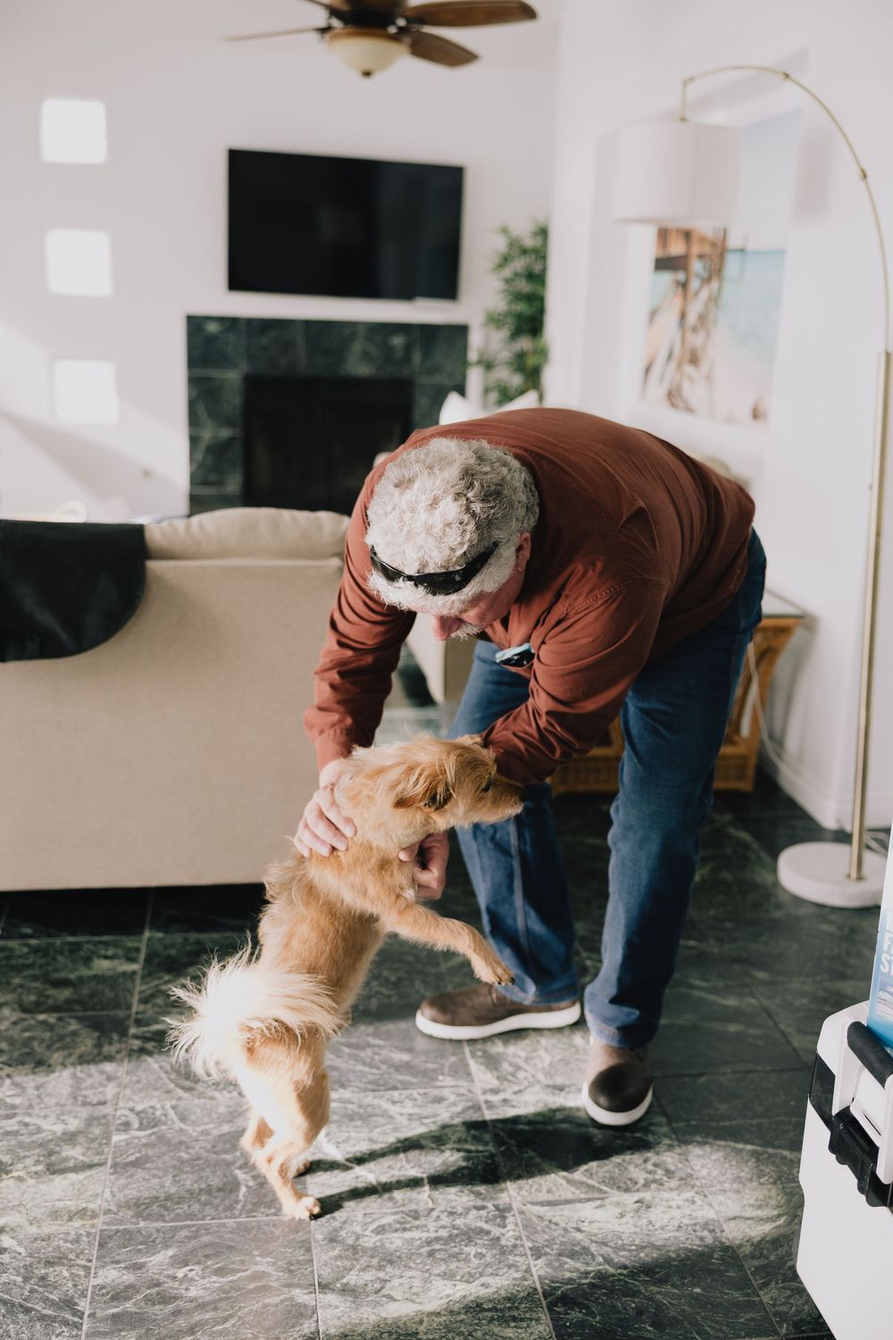 Senior man playing with small golden dog in modern living room with marble floors.
