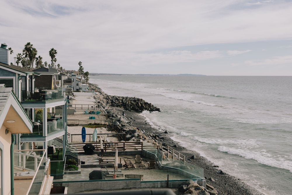 Coastal beachfront homes with balconies overlook a rocky shoreline and calm ocean waves under an overcast sky.