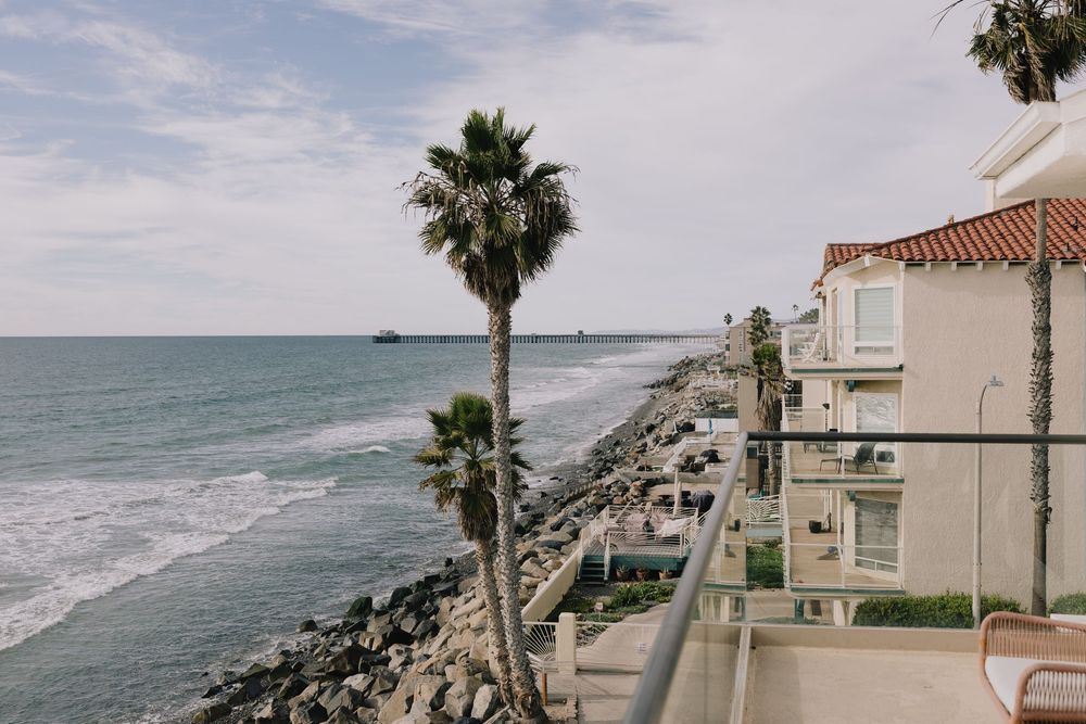 Oceanfront balcony view with palm tree, rocky shoreline, and coastal buildings under cloudy skies.