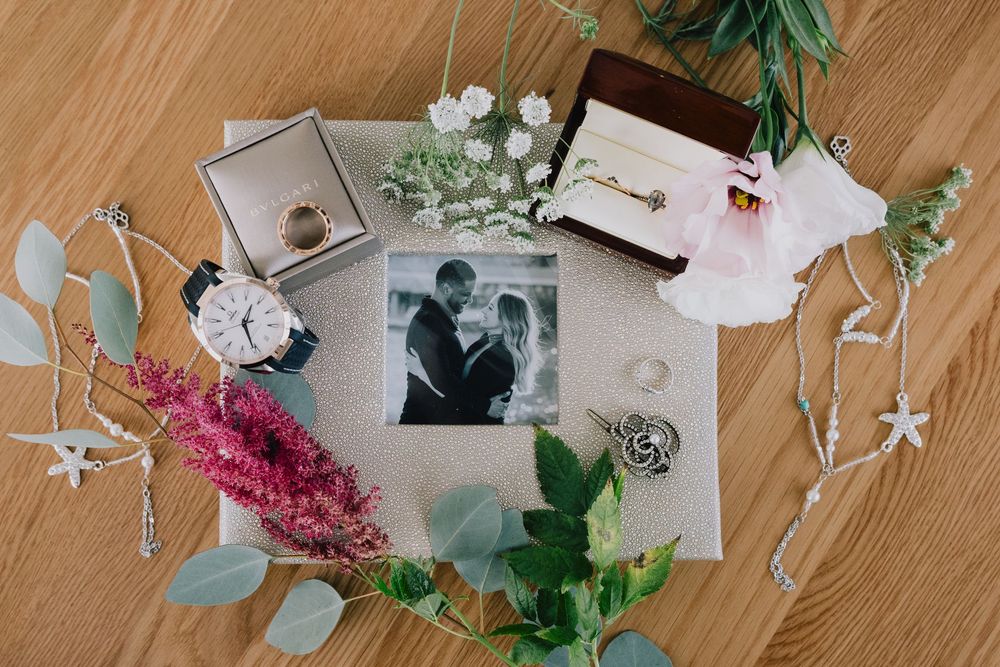 Wedding flat lay with rings, watch, flowers, and black and white couple photo on wood surface.