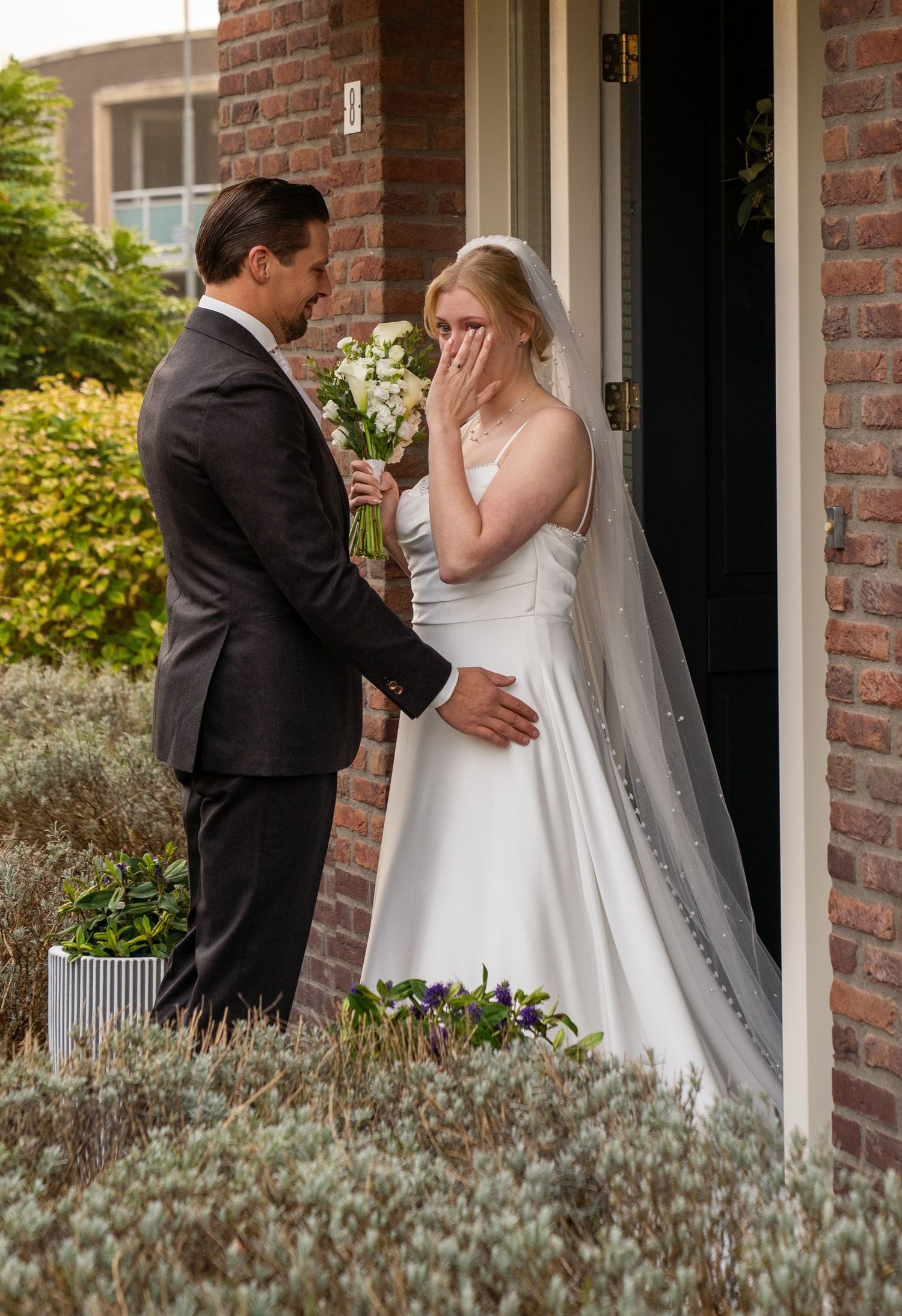 Een geëmotioneerde bruid bedekt haar gezicht terwijl de bruidegom het boeket overhandigt tijdens de 'first look' in het bakstenen huis.