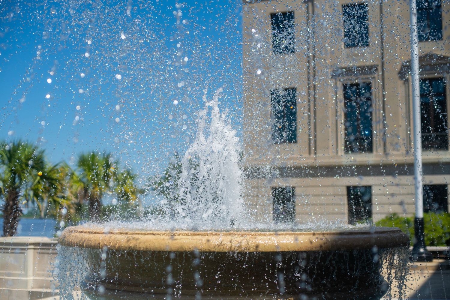 A decorative stone fountain sprays water in front of a classical building with palm trees under a clear blue sky.