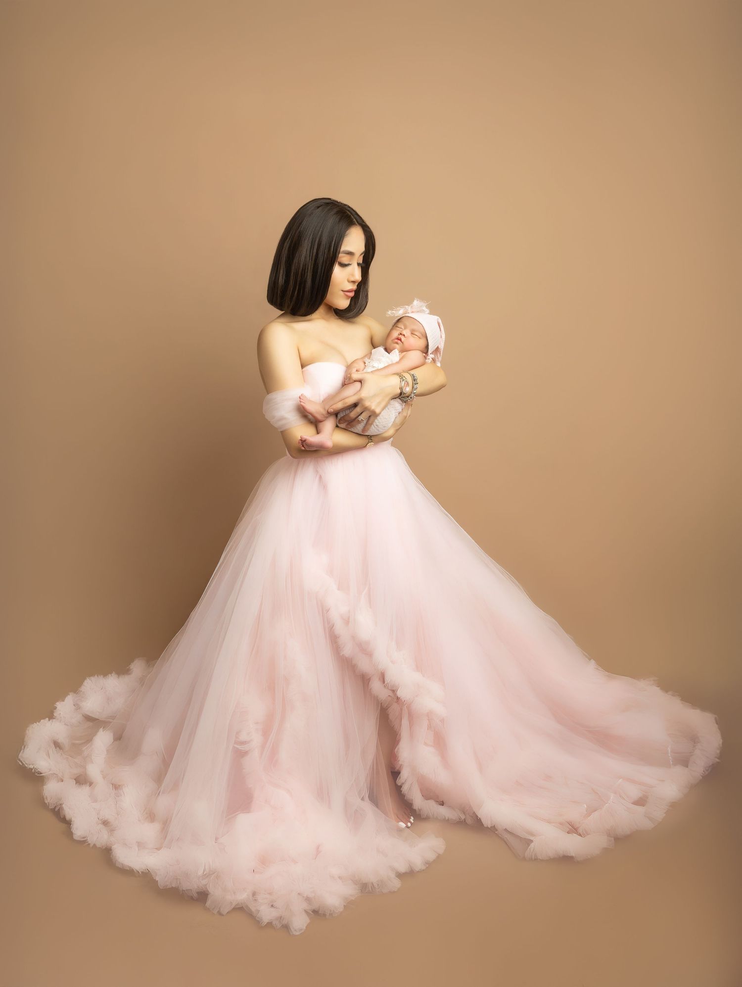 A woman in a pink tulle gown holds a sleeping newborn baby in a professional studio portrait.