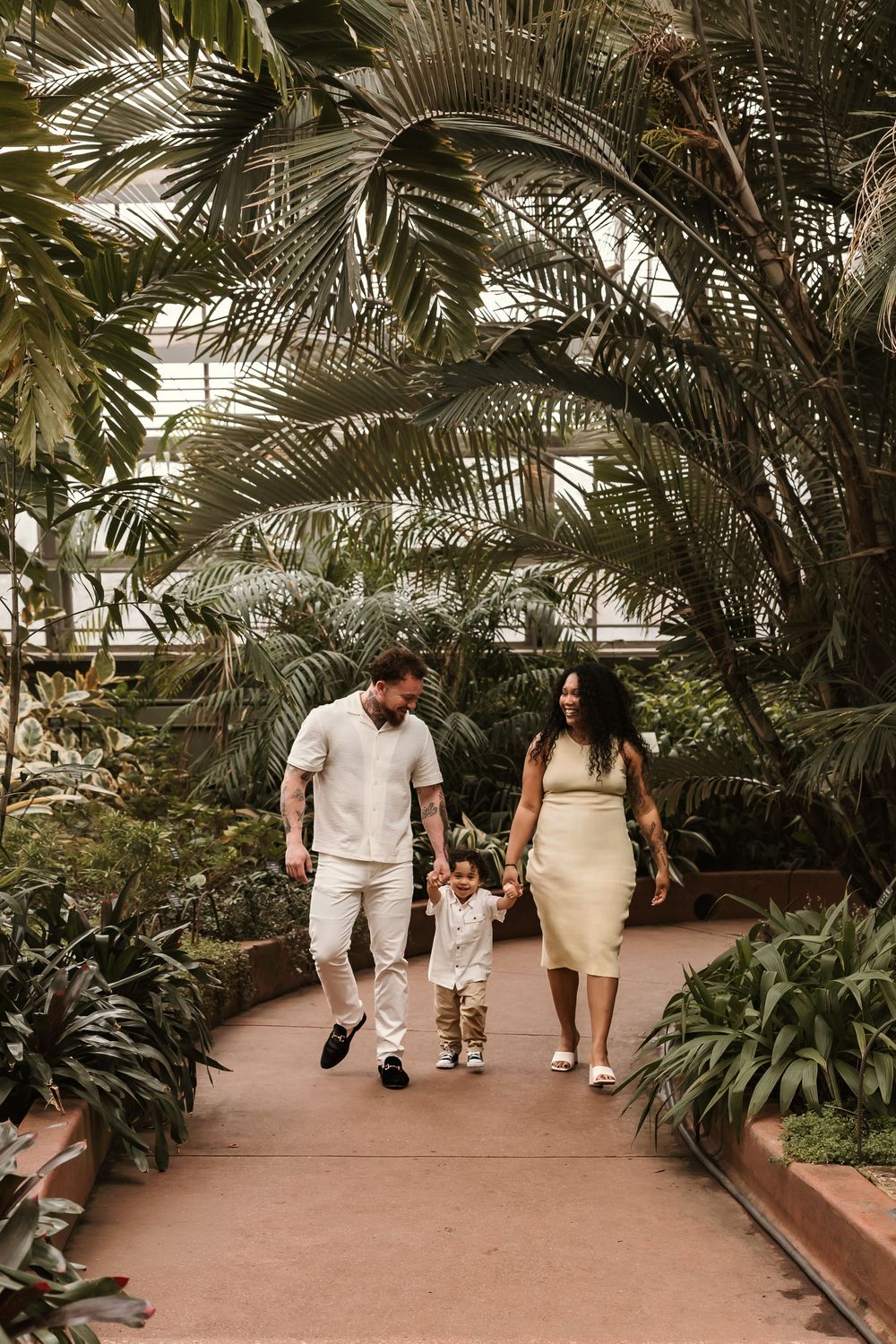Happy interracial family of three walks hand-in-hand through lush tropical greenhouse garden pathway.