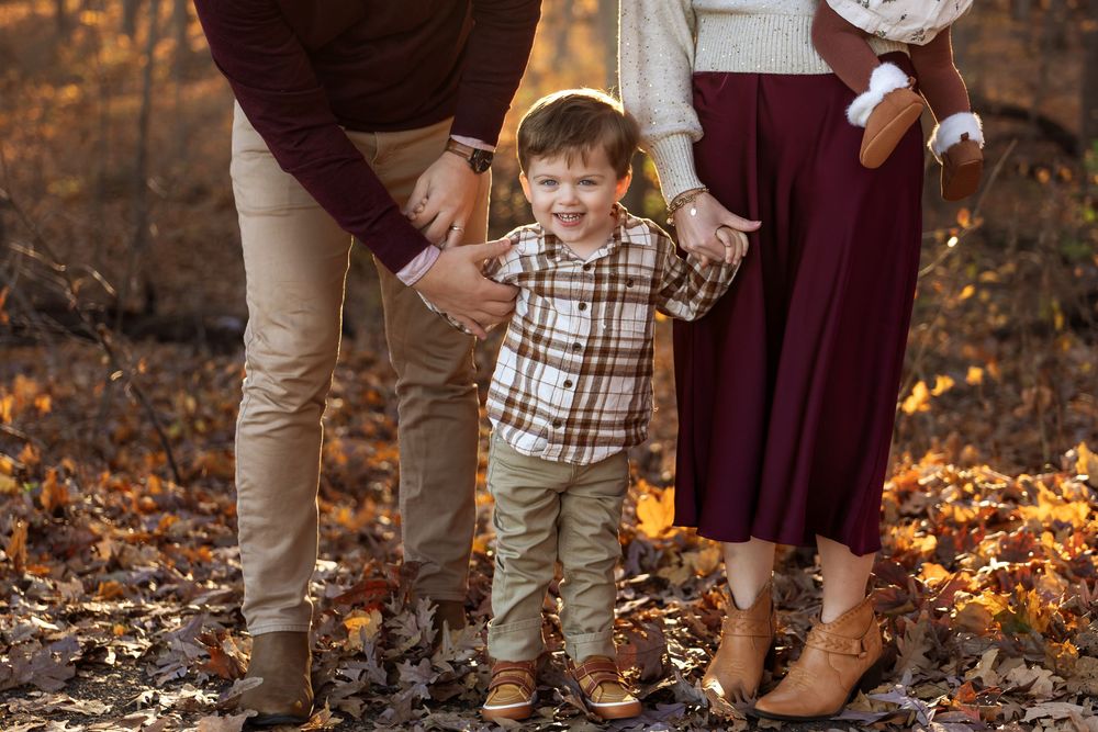 Parents hold hands with young toddler boy in plaid shirt during fall family photo session.
