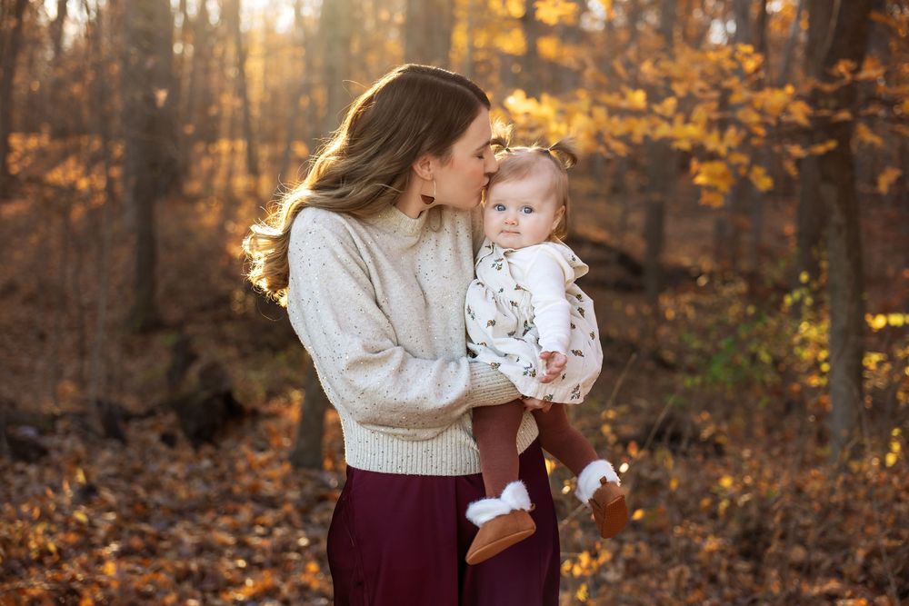Mother in cream sweater kisses baby girl while holding her during golden hour fall portrait.