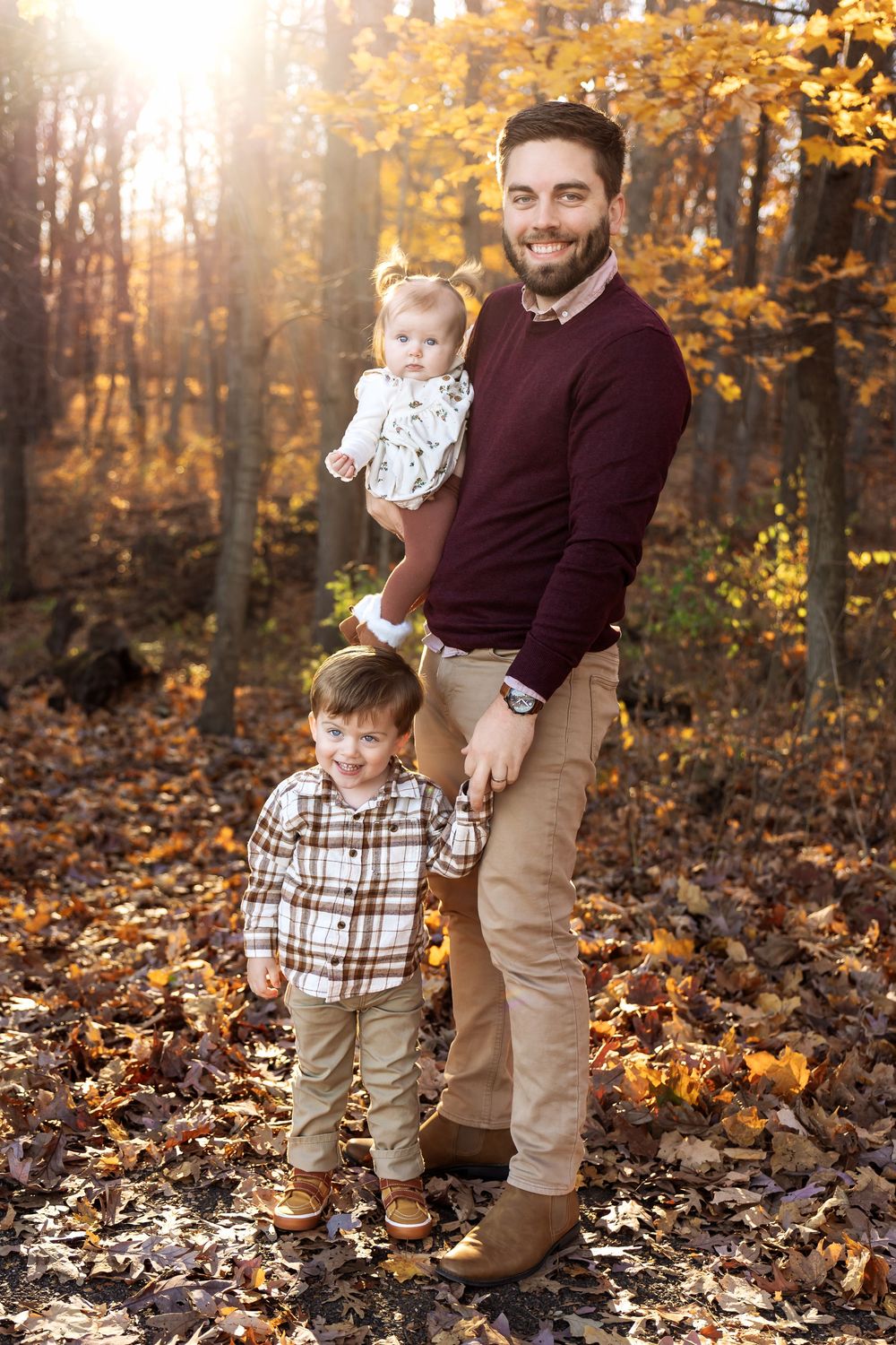 Smiling bearded father holds infant daughter while young son stands beside him in autumn forest.