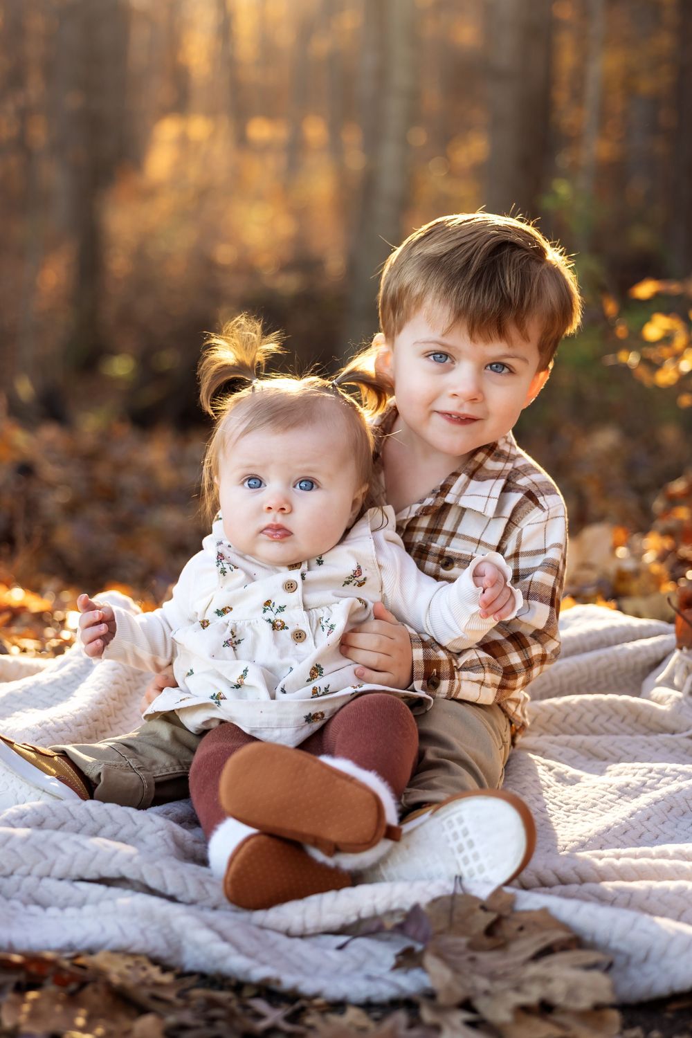 Two young siblings with blue eyes sit together on a blanket in an autumn forest during golden hour.