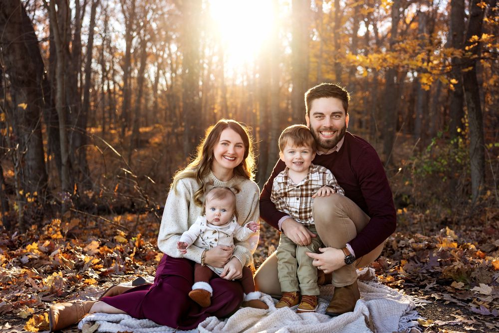 Young family of four poses on blanket in golden autumn forest, parents holding two small children.
