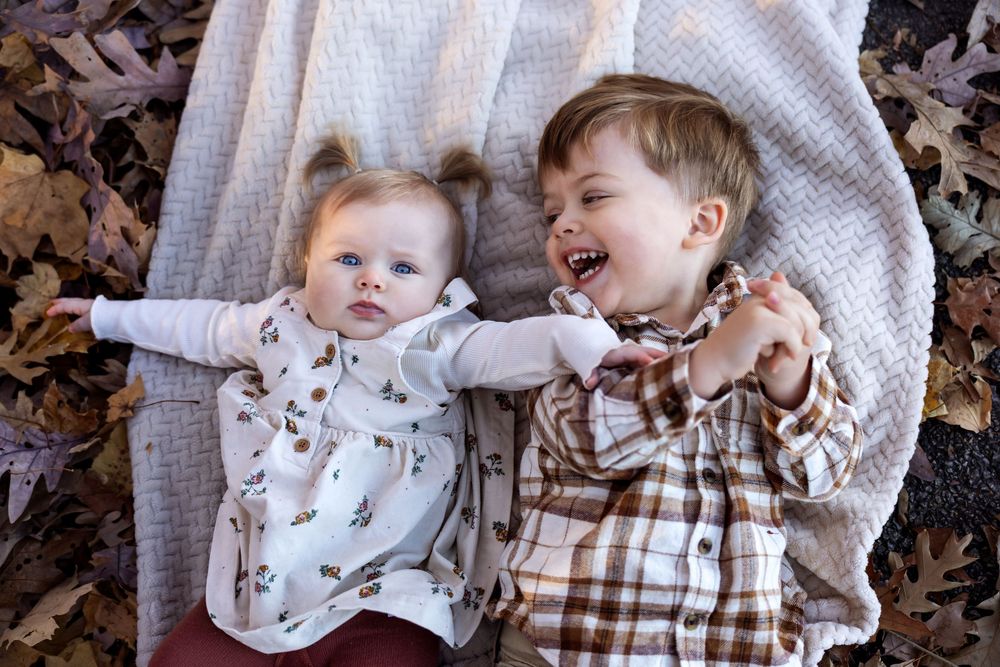 Baby girl and toddler boy laugh together lying on blanket surrounded by colorful autumn leaves.