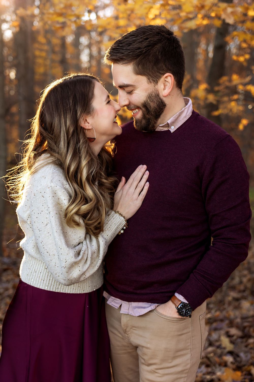 Romantic couple nearly kissing in warm autumn forest light, woman in cream sweater and burgundy skirt.