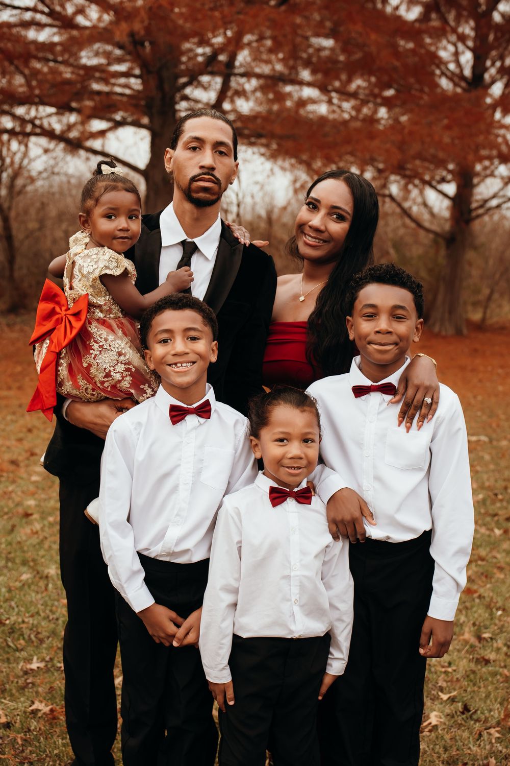 Elegant family of six in formal red and black attire poses together in colorful autumn park setting.