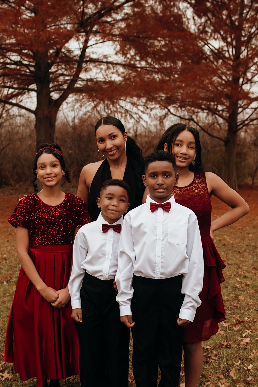 Mother in black dress poses with four children in red and white formal attire in autumn park.
