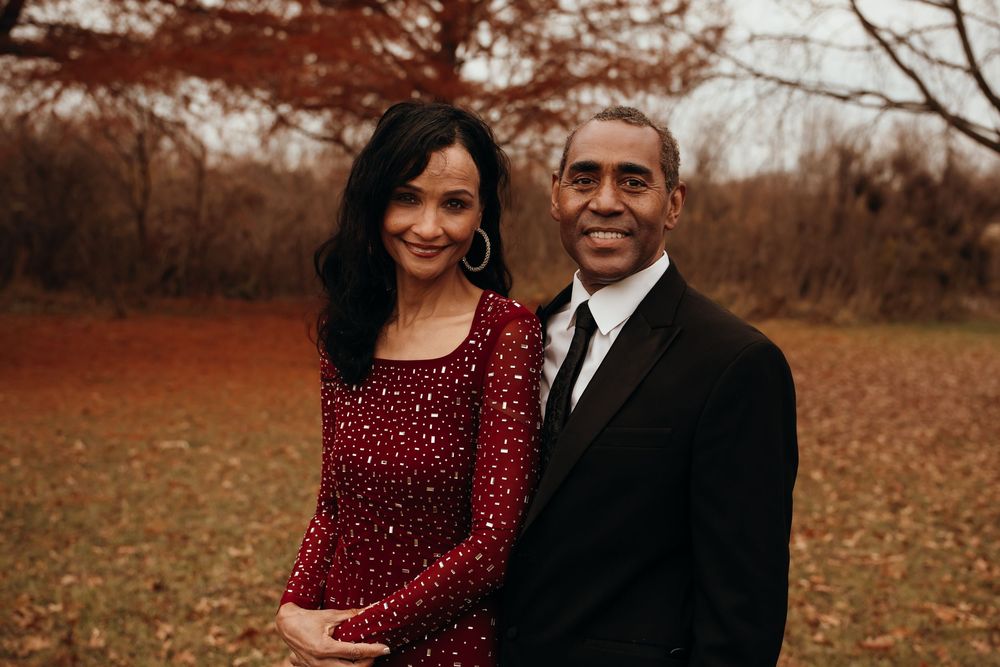 Smiling couple in formal red and black attire embraces warmly outdoors in autumn landscape setting.