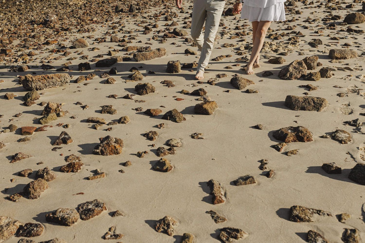 Detail of couple walking barefoot on sandy beach with rocks during engagement session