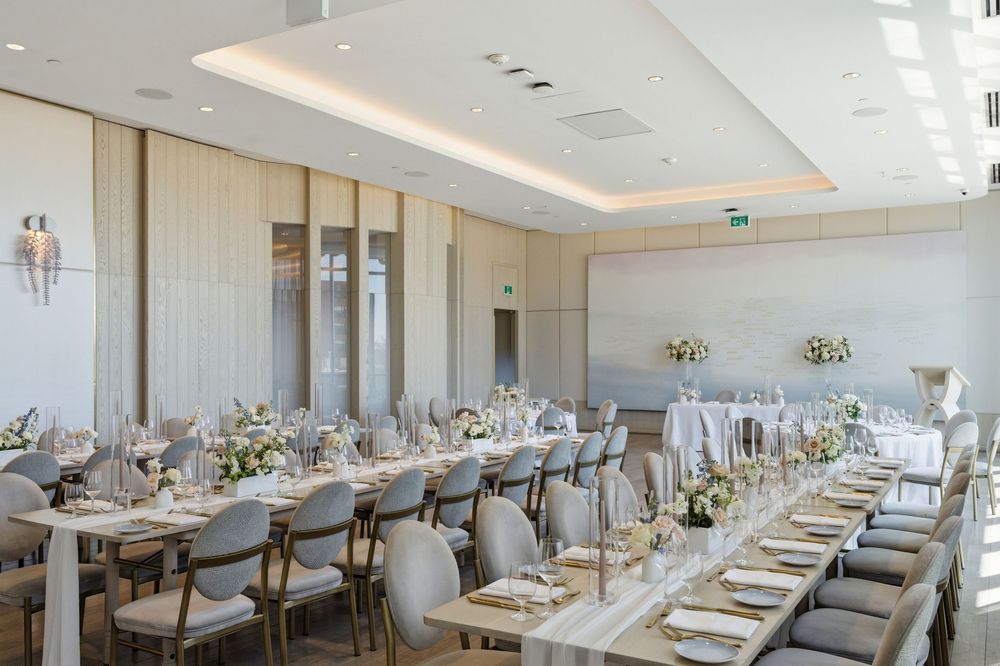 Elegant banquet hall with long tables set with white linens and gold chairs for a formal wedding reception.