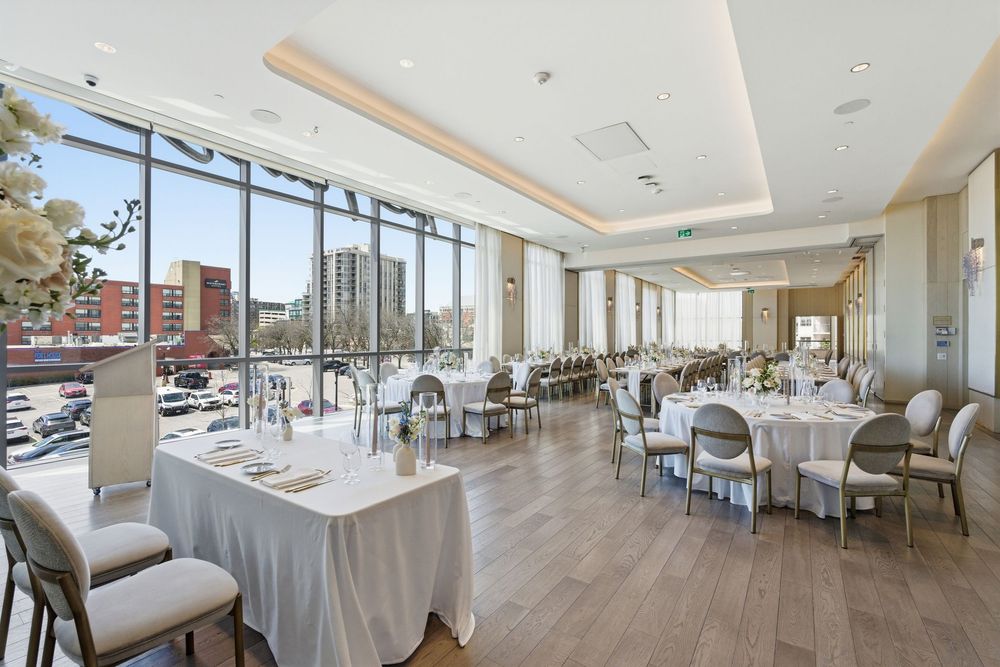 Elegant banquet hall with floor-to-ceiling windows, white tablecloths, and modern decor overlooking a city view.
