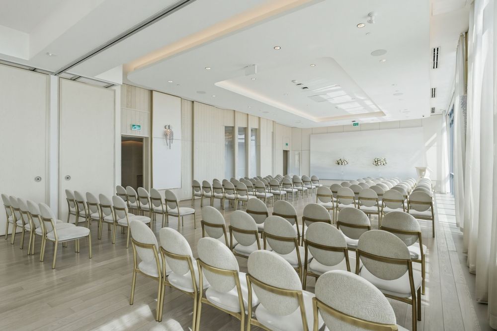Conference room setup with rows of white chairs and natural lighting from large windows.