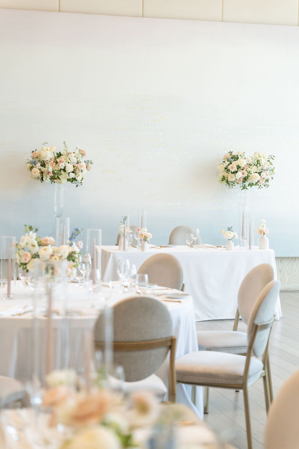 Round dining tables with white tablecloths and beige chairs decorated with delicate floral centerpieces in a light-filled room.
