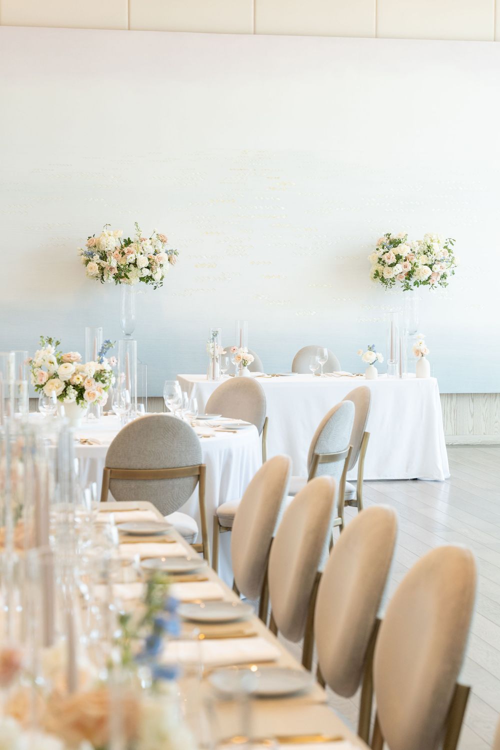 Elegant wedding reception table setting with white linens, beige chairs, and delicate floral centerpieces in glass vases.