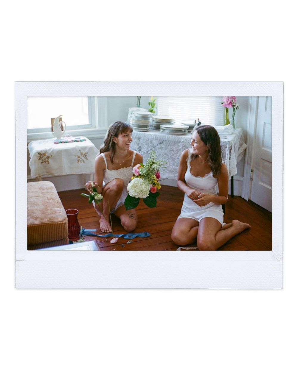 A film photo of bride and friend help arrange flowers sitting on a wooden plank floor in a sunny apartment in Warren, Rhode Island