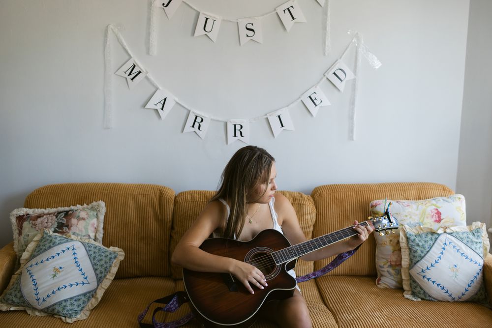 A young woman sits on mustard colored couch with pillows on it with a Just Married sign above her playing a guitar.. Warren RI wedding photo
