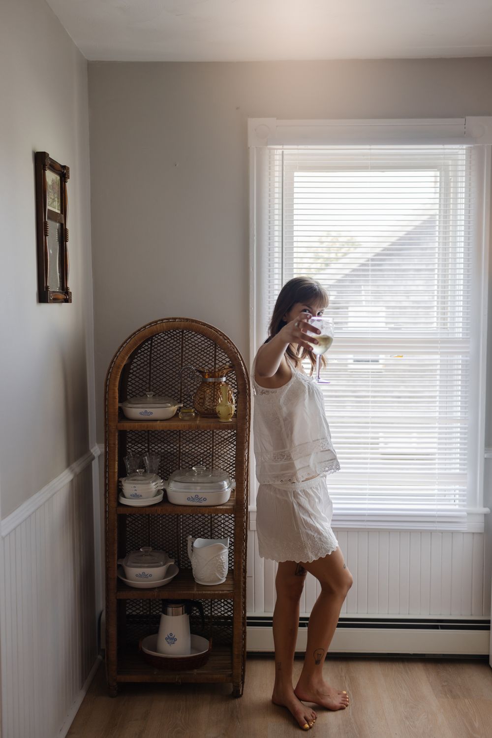 Bride wears cotton shorts and tank pajamas stands next to window and wicker shelf toasting photographer with a wine glass. Warren RI wedding