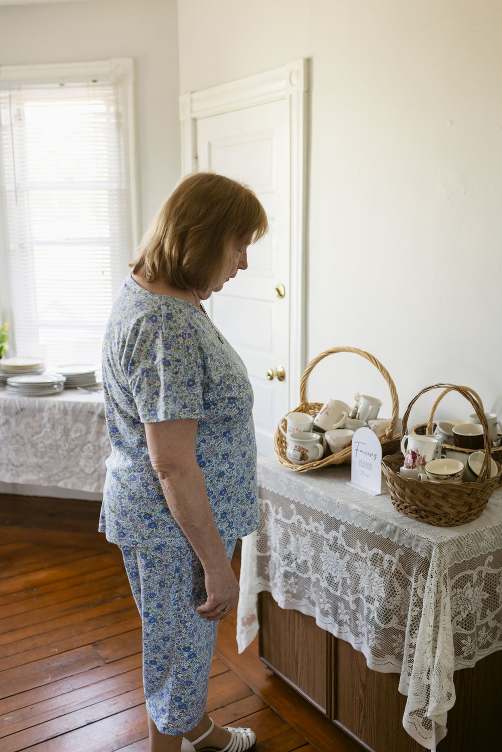 Older woman in blue pajamas stands in a bright living room looking at vintage coffee mugs and teacups in a wicker basket in Warren RI.