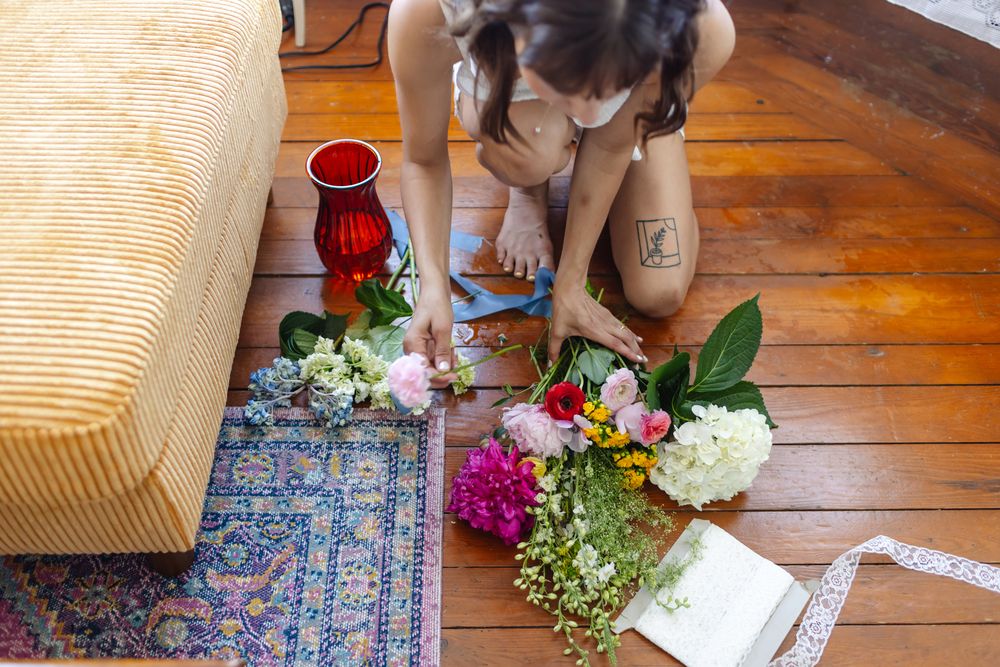 Bride puts together bouquets on wooden plank floor next to yellow couch. York, Cohasset, Newport, Essex County wedding photographer