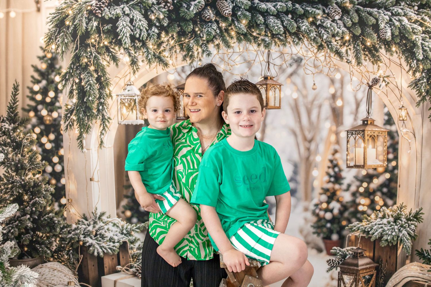 Mother holding two children in green outfits in festive Christmas setting.