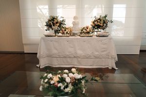 Elegant white wedding cake with cascading fresh flowers displayed on glass table in bright reception venue.