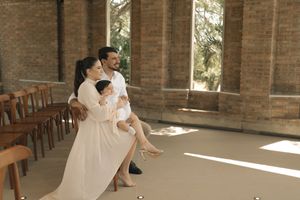 A couple in white attire sits together in a sunlit church interior with wooden pews and brick walls.