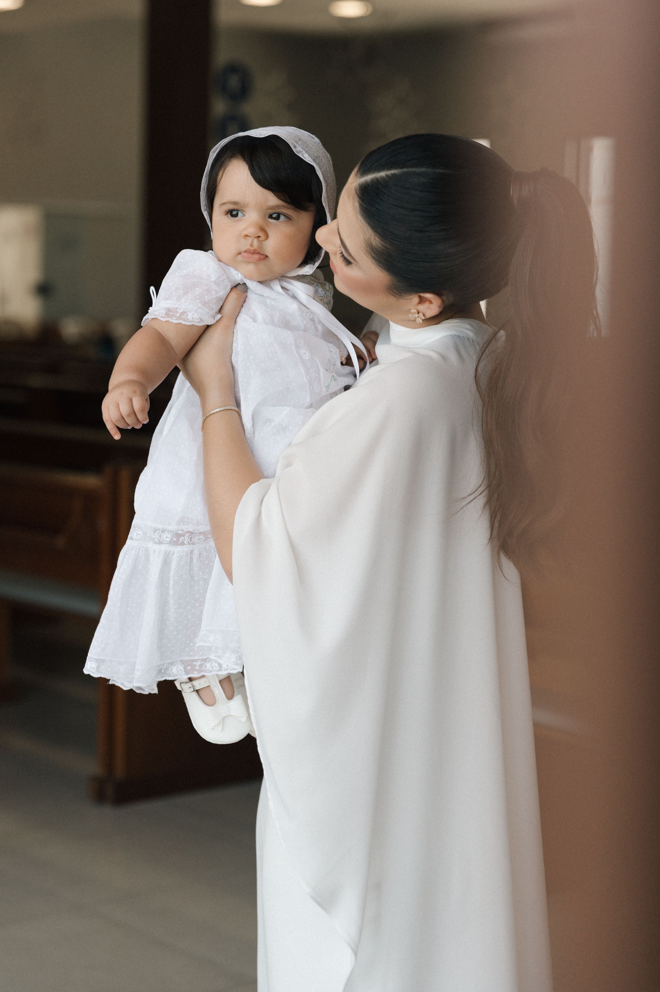 A tender moment captured in white as a parent in flowing robes holds a baby wearing a delicate white dress and headband.