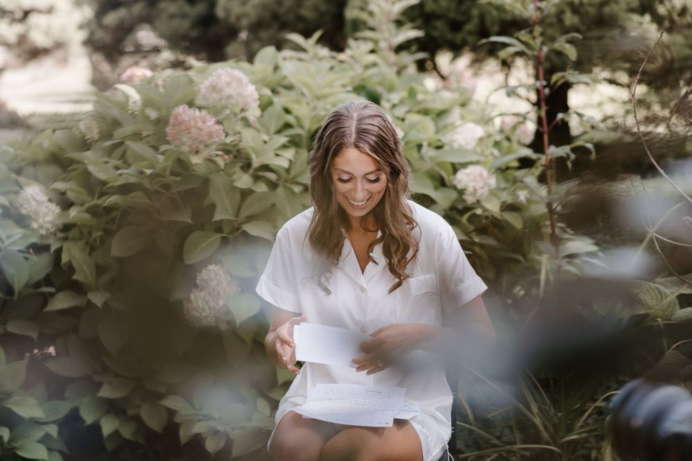 Bride in white robe reading a letter from groom surrounded by blooming hydrangeas outdoors.