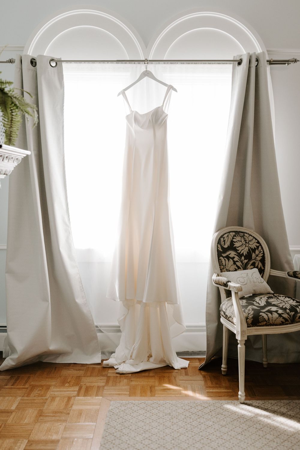 Simple elegant white mermaid wedding dress hanging in sunlit window beside vintage French chair.