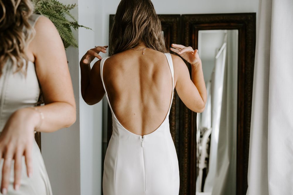 Bride in backless white wedding gown being helped by bridesmaid in front of mirror.