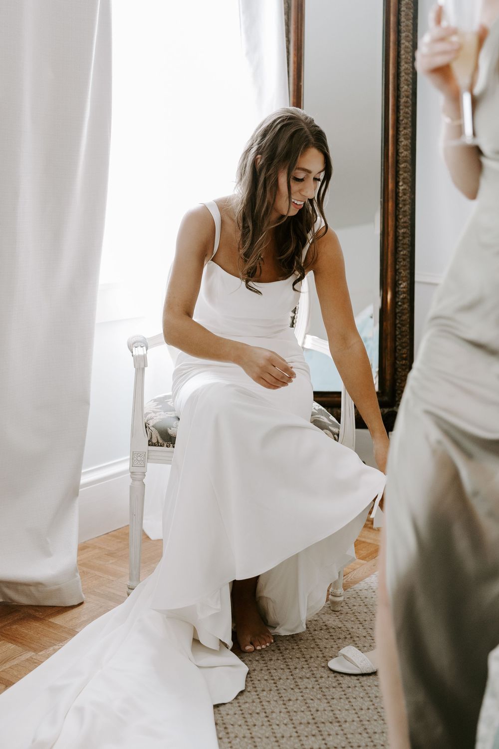Bride in elegant white wedding dress sitting in chair adjusting her shoes before ceremony.